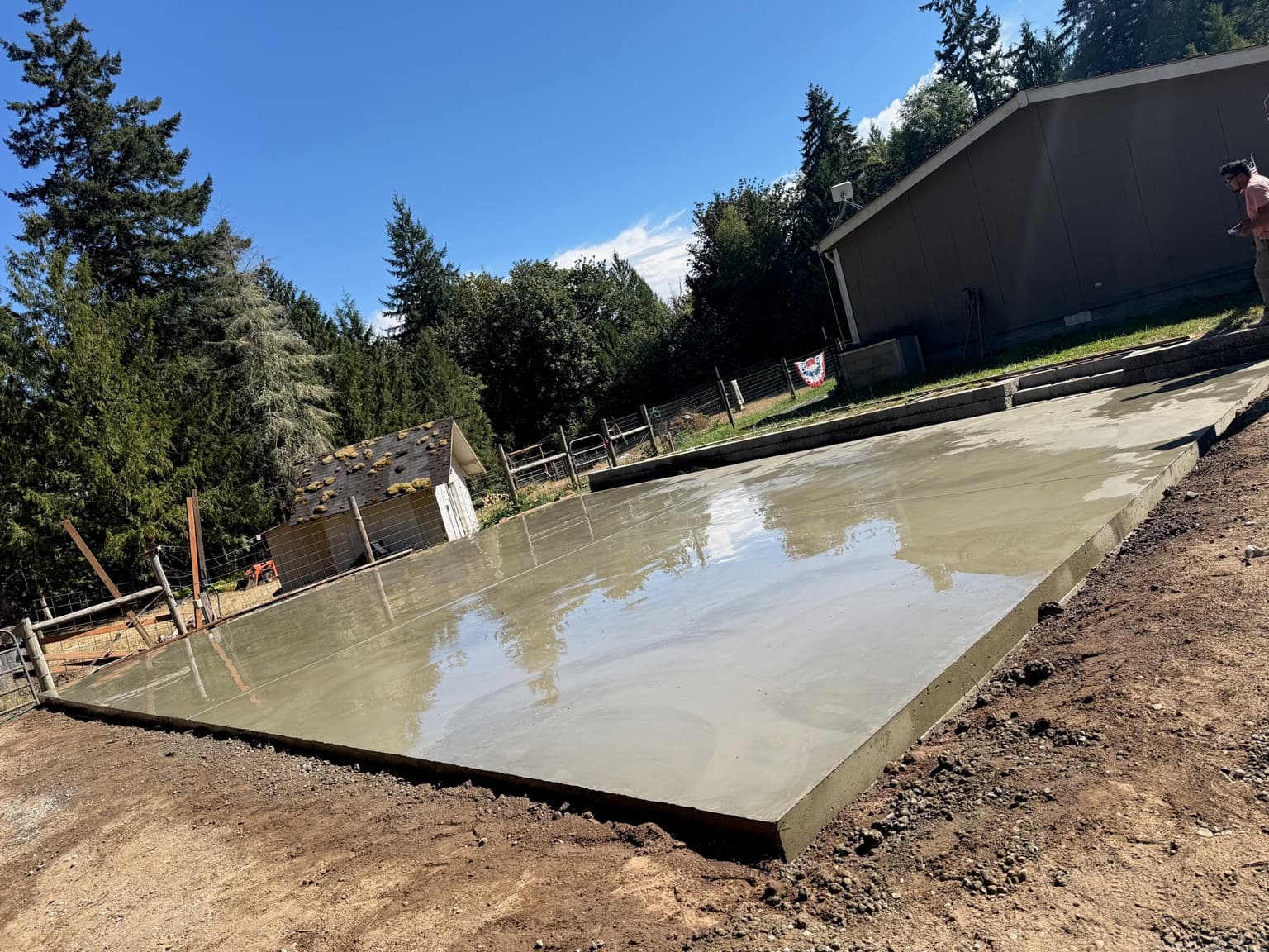 Freshly poured and float-finished concrete slab beside an outbuilding, edges formed and cut clean